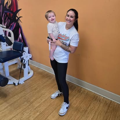 Smiling member holding a toddler beside a training machine and wall mural