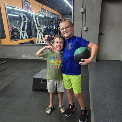 Two smiling young boys on the gym floor, one holding a medicine ball